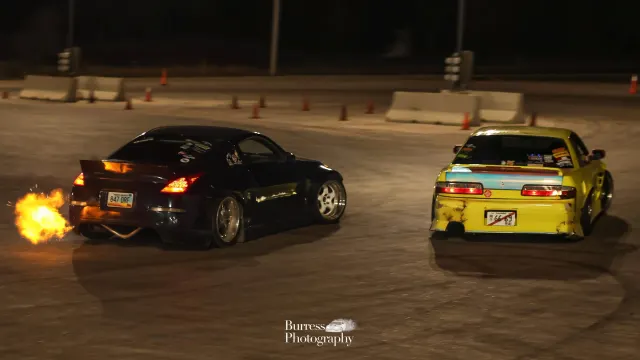 Two cars mid-drift at night at the Thunderdome, one throwing flames from the exhaust